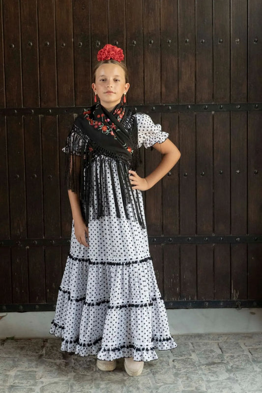 Young girl in a traditional dress with a black and white pattern, standing against a wooden door.