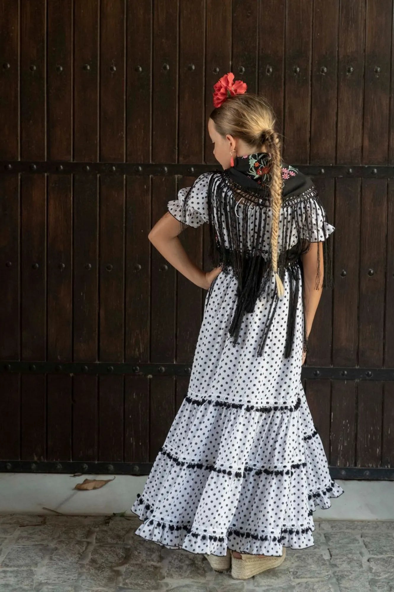 Niña con vestido de lunares blanco y negro, con una trenza larga y una flor roja, frente a una puerta de madera.