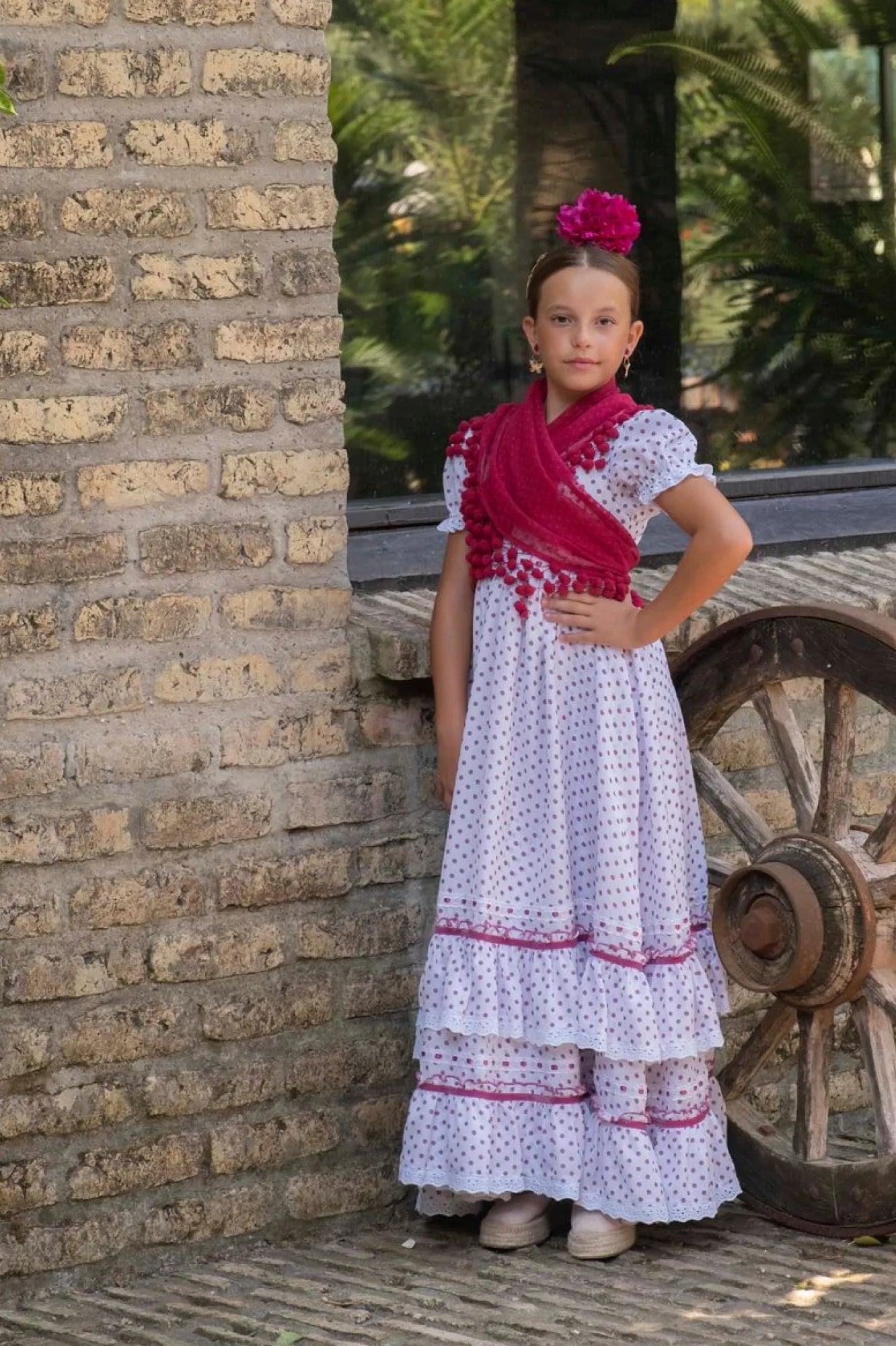 Young girl in a traditional outfit standing next to a brick wall and old wheel.