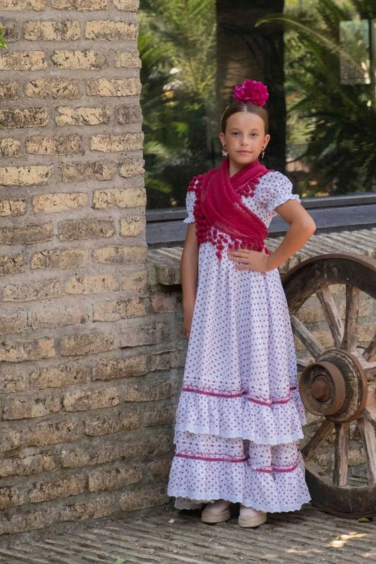 Young girl in a traditional outfit standing next to a brick wall and old wheel.