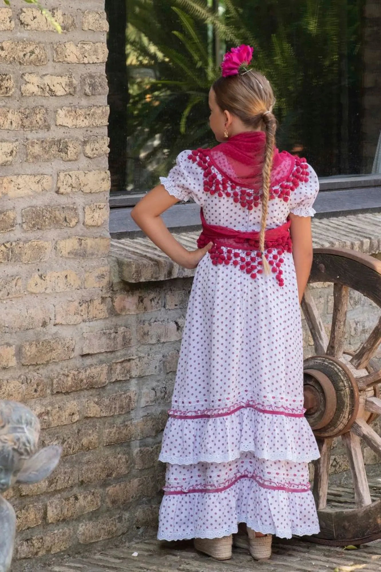 Woman in a white dress with red patterns standing next to a stone wall and wooden cart wheel.